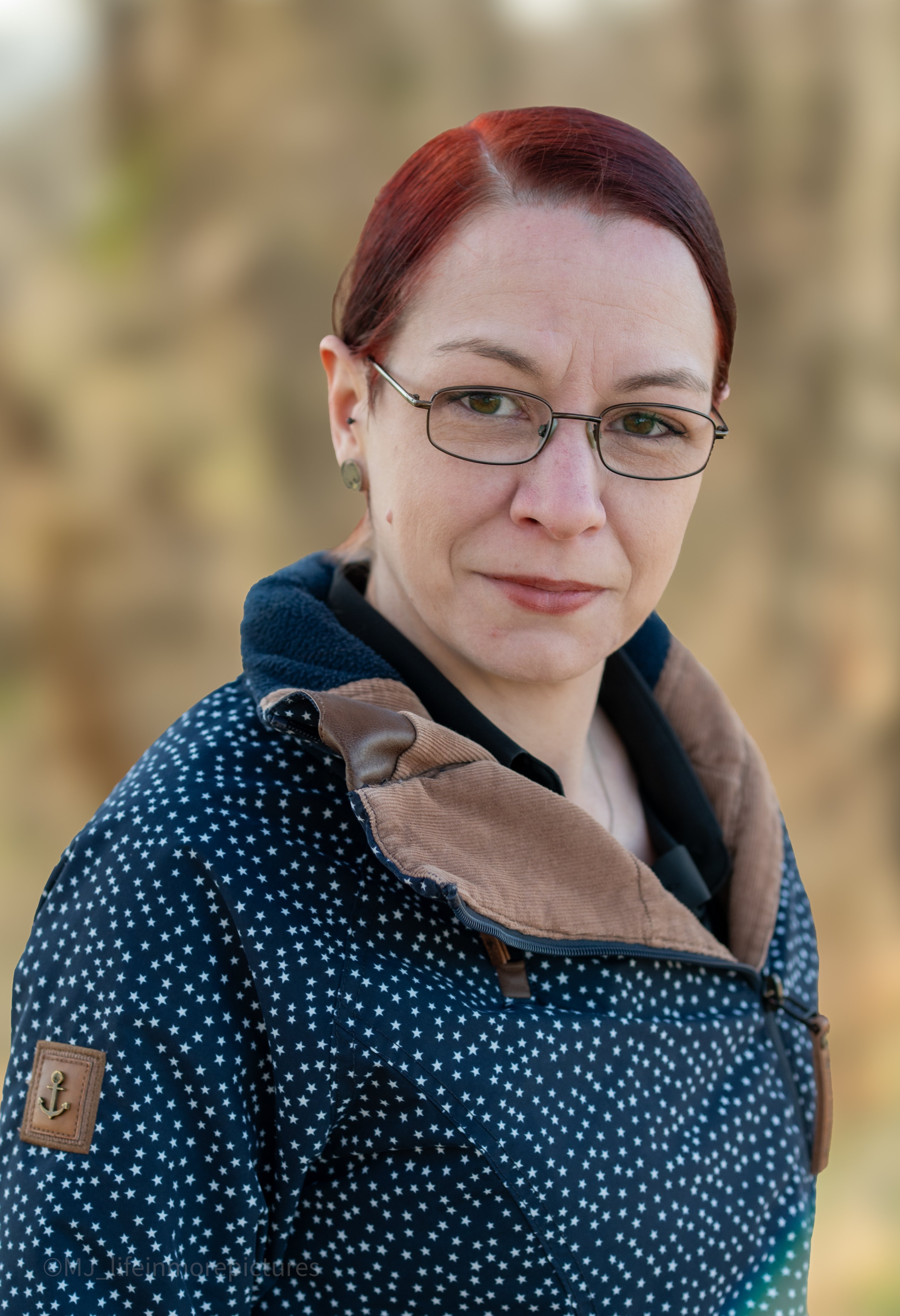 Lächelnde Frau mit Brille und rotem Haar Das Bild zeigt eine Frau mittleren Alters, die in einer natürlichen Umgebung steht und freundlich in die Kamera lächelt. Sie trägt eine Brille mit schwarzem Gestell und hat rote Haare, die zu einem Pferdeschwanz zusammengebunden sind. Ihre Kleidung besteht aus einem dunkelblau-weiß gemusterten Mantel, der ihre Figur umspielt. Insgesamt vermittelt das Bild einen warmherzigen und offenen Eindruck von der abgebildeten Person.