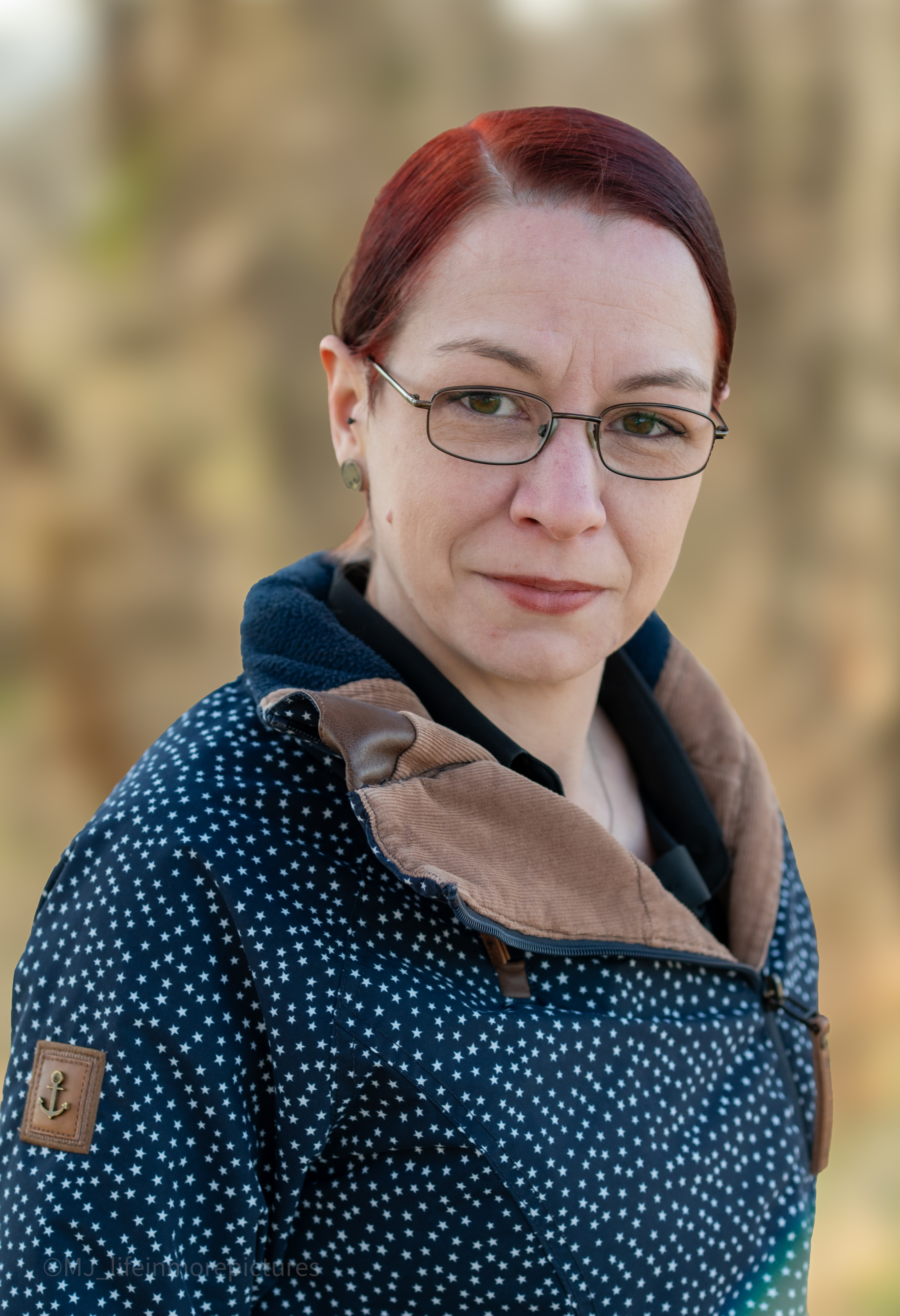 Lächelnde Frau mit Brille und rotem Haar Das Bild zeigt eine Frau mittleren Alters, die in einer natürlichen Umgebung steht und freundlich in die Kamera lächelt. Sie trägt eine Brille mit schwarzem Gestell und hat rote Haare, die zu einem Pferdeschwanz zusammengebunden sind. Ihre Kleidung besteht aus einem dunkelblau-weiß gemusterten Mantel, der ihre Figur umspielt. Insgesamt vermittelt das Bild einen warmherzigen und offenen Eindruck von der abgebildeten Person.